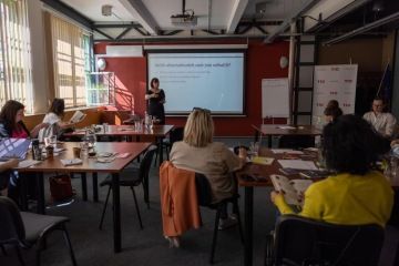 Skupina lidí sedících kolem stolu během workshopu v moderní zasedací místnosti. Facilitátorka stojí před plátnem s projekcí, na které je zadání řešené designové výzvy.