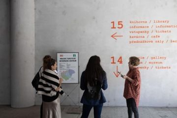 Tři ženy stojí u stěny s orientačními nápisy v turistickém informačním centru. Na stěně jsou směrovky k různým sekcím centra, jako knihovna, galerie a kavárna, napsané v češtině a angličtině.